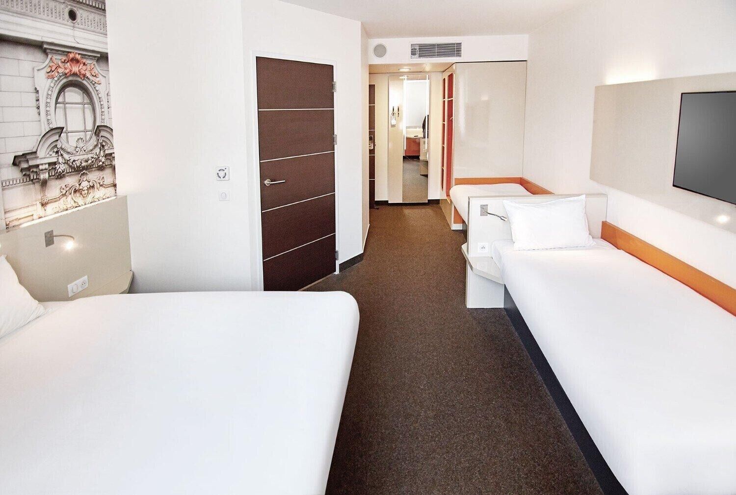A hotel room with two beds, a wardrobe, a television, and a hallway leading to another room.