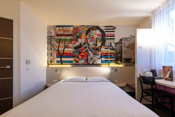 A hotel room with a bed, a desk, and a large colorful mural on the wall.