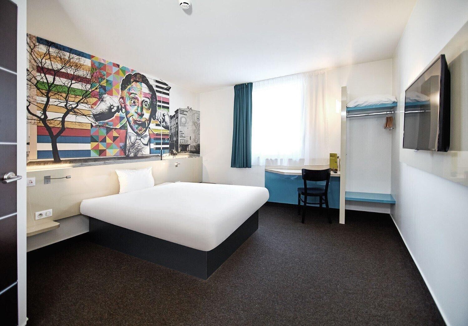 A hotel room with a large bed, a desk, a chair, a TV, and a colorful mural on the wall.