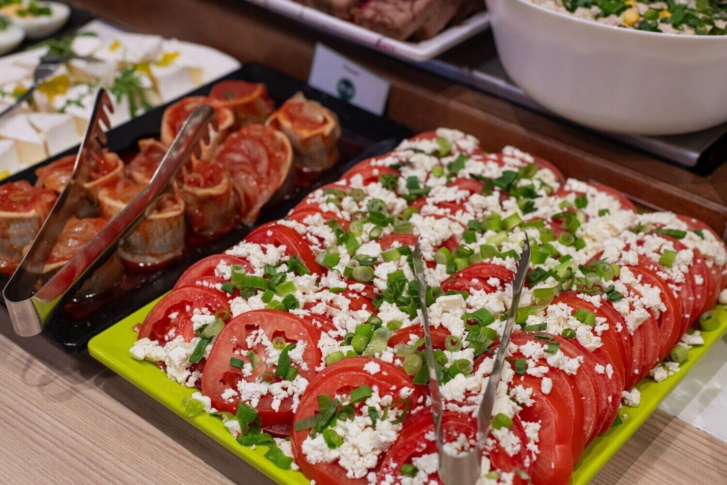 Een buffet met diverse gerechten, waaronder een groot schaal met plakjes tomaten en feta, versierd met groene bieslook.