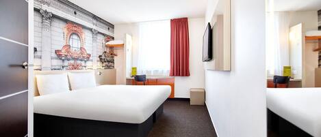 Ein Hotelzimmer mit einem großen Bett, einem Schreibtisch, einem Sessel, einem kleinen Tisch, einem Spiegel, einem Fenster mit Vorhängen und einer Wand mit Wandmalerei.