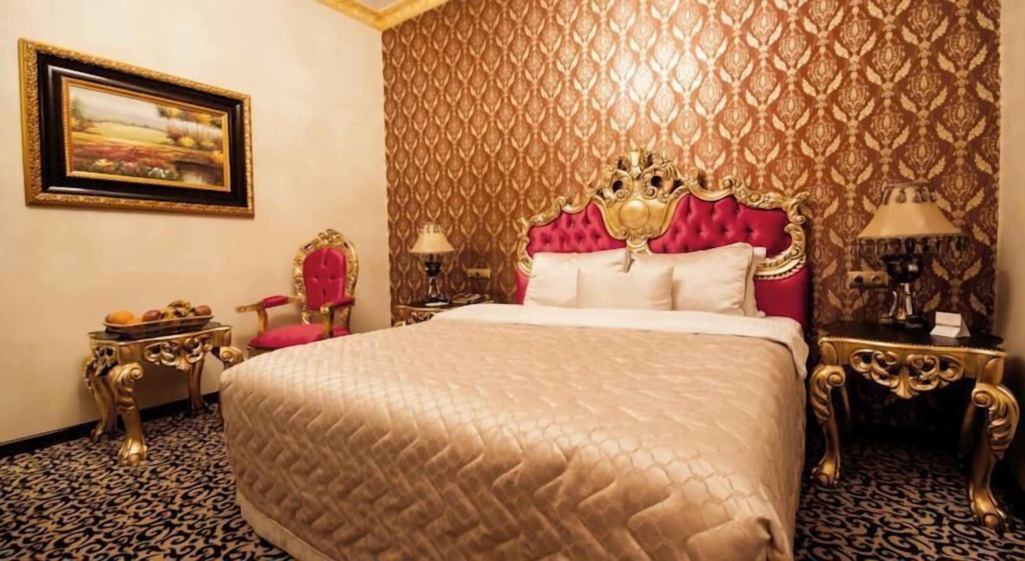 A luxurious hotel room with a large bed, ornate headboard, and a patterned carpet.