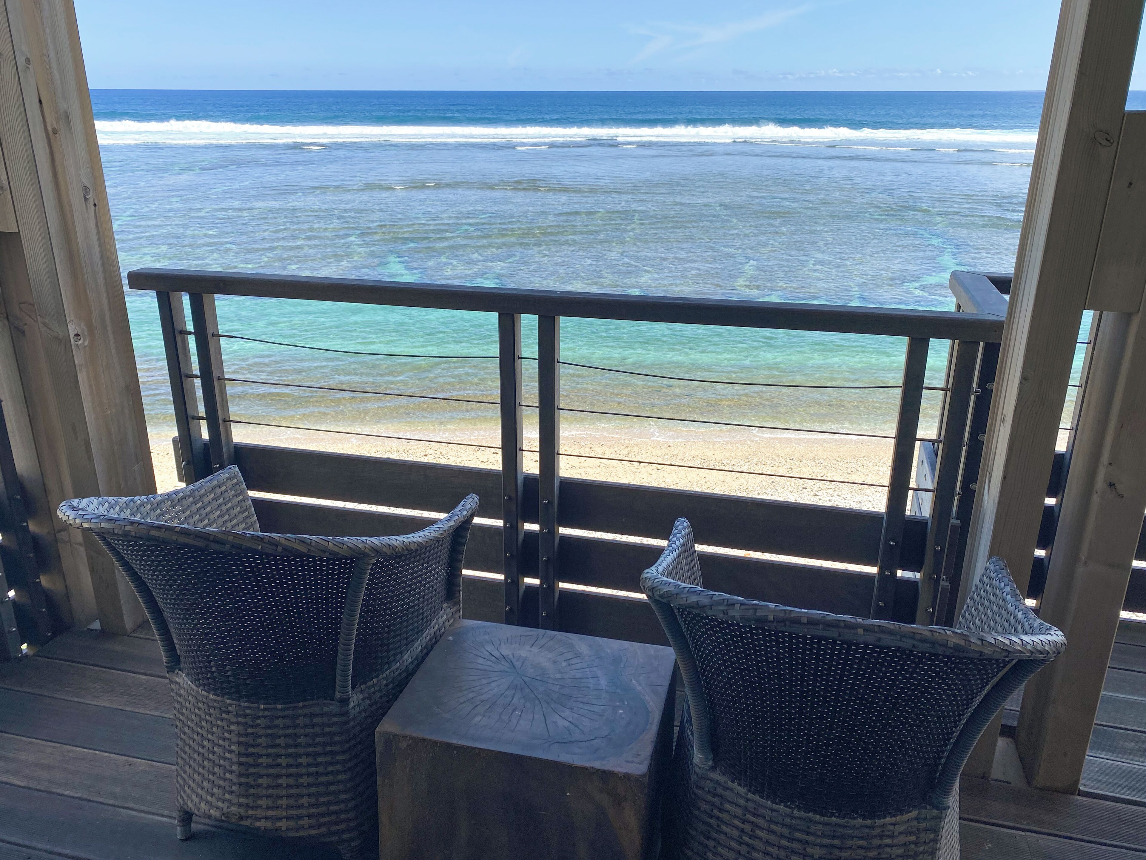 A wooden deck with wicker chairs and a table overlooking a beach with clear turquoise water and waves.