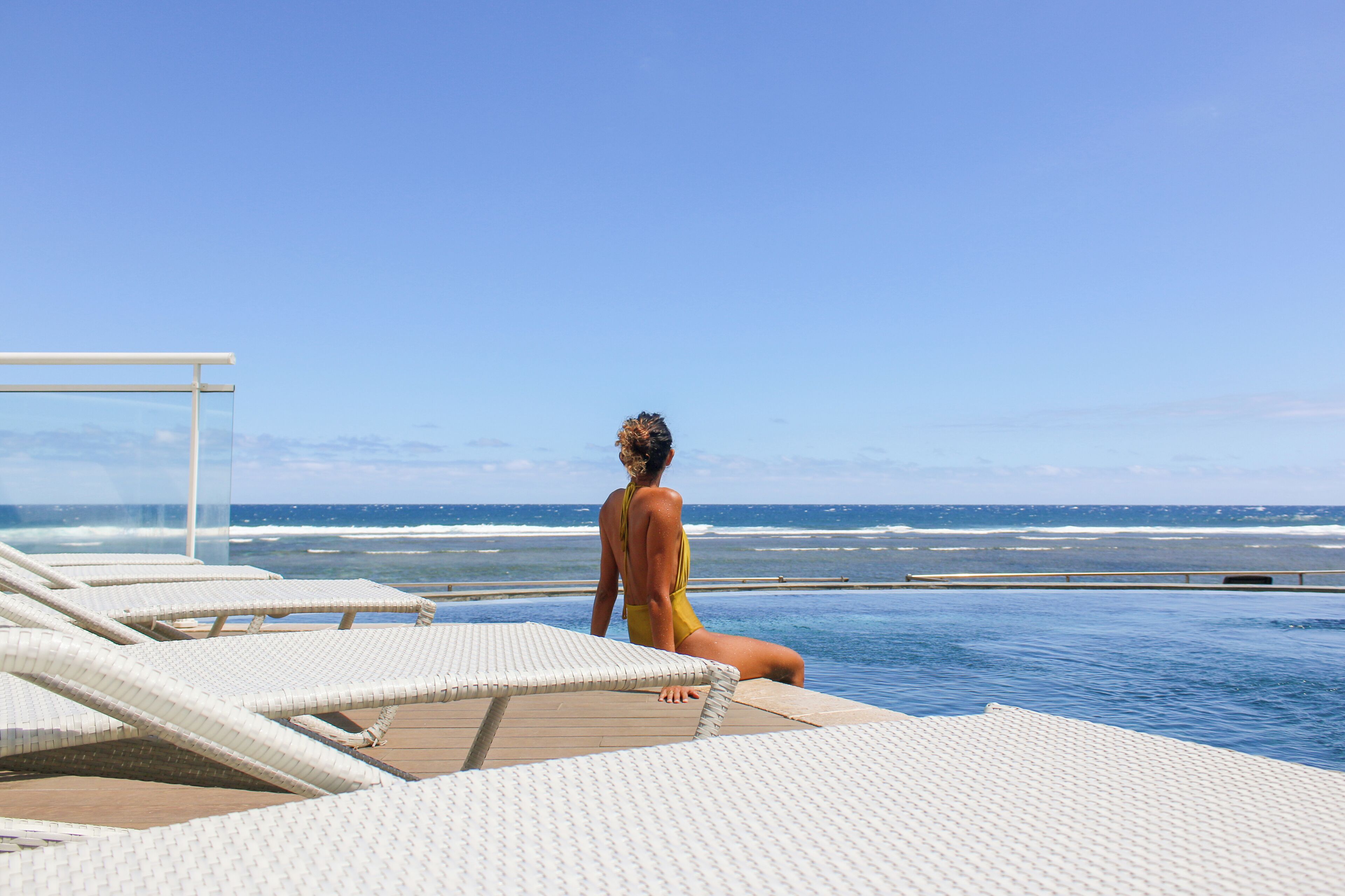 Una mujer con traje de baño amarillo sentada en una silla lounge junto a la piscina, con vista al océano.