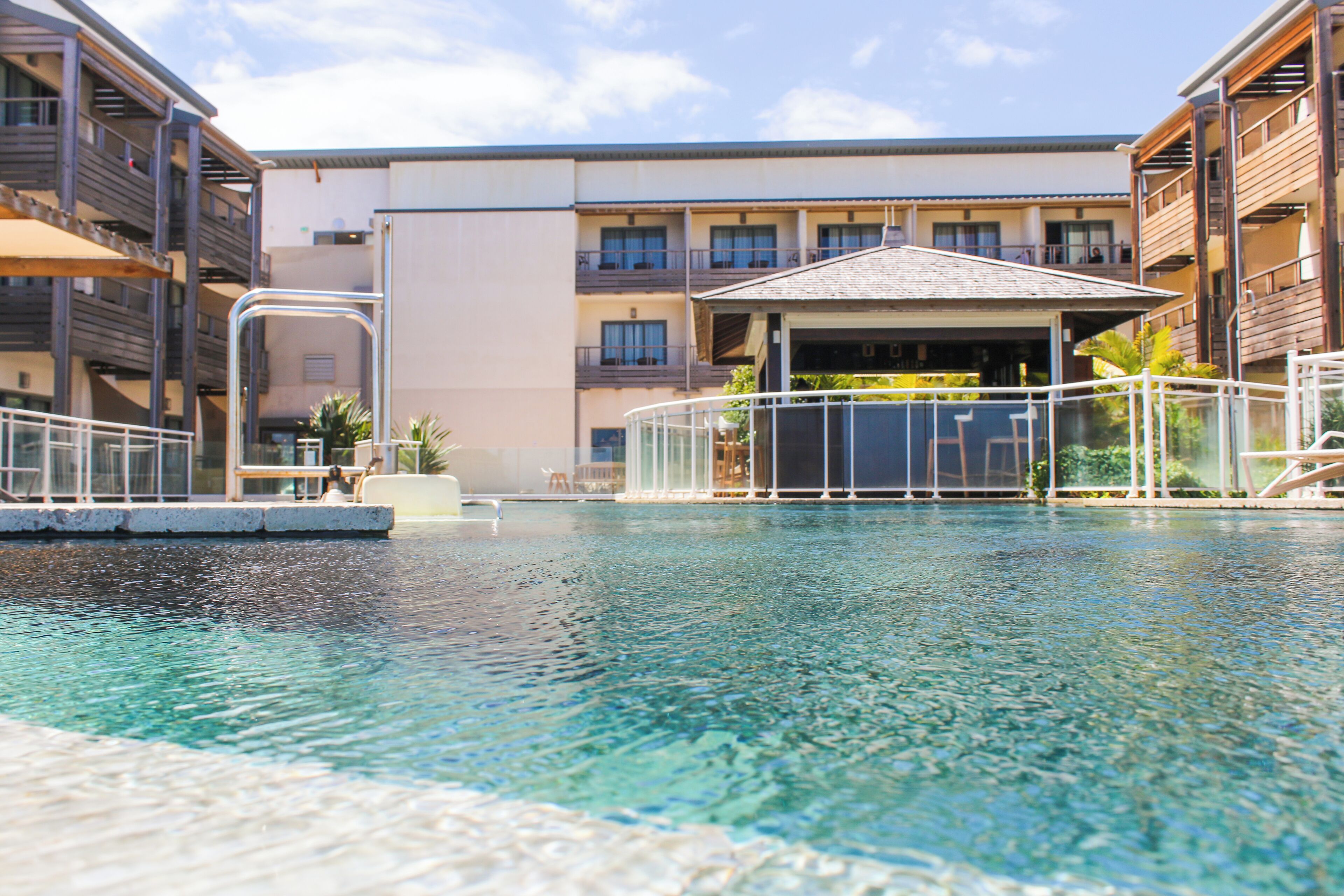 A swimming pool with a clear blue water surface, surrounded by a white poolside area and a building with multiple balconies and a covered patio area.