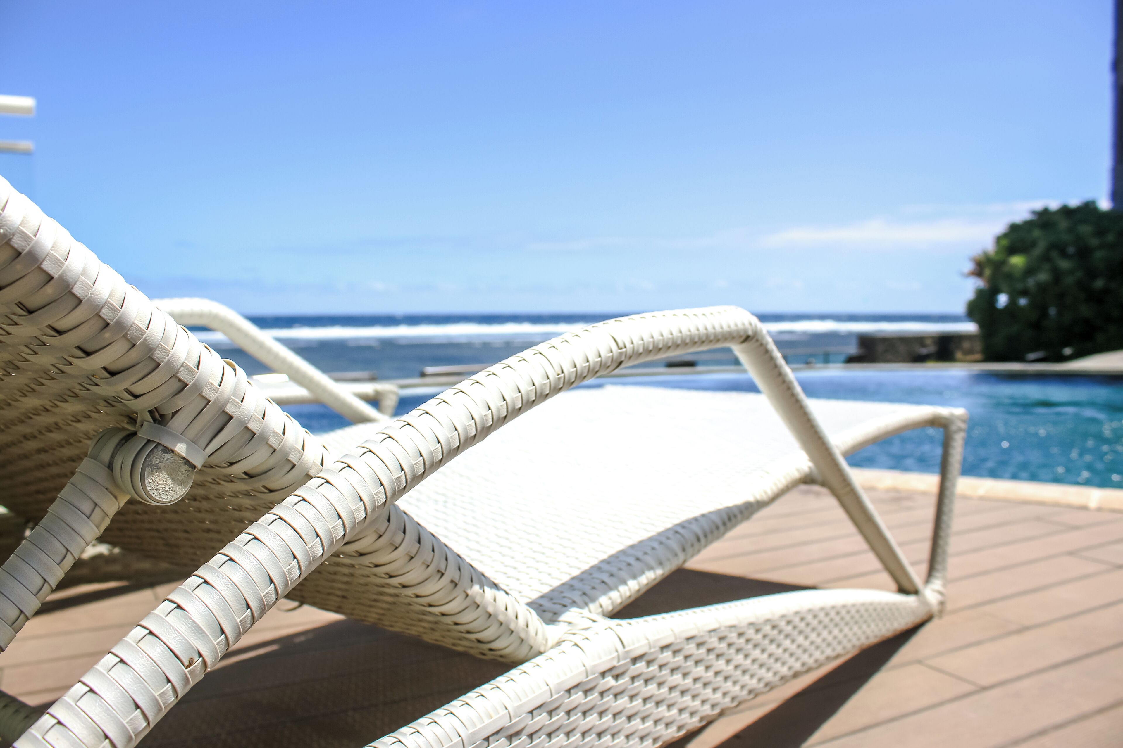A white wicker lounge chair by a pool with a view of the ocean.