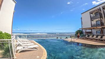 A pool with clear blue water, a wooden deck, and lounge chairs. In the background, there is a view of the ocean and a building with balconies.