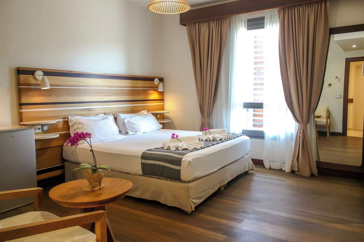 Une chambre d’hôtel avec un grand lit, des tables de chevet, un petit bureau, une chaise, un plancher en bois et une fenêtre avec des rideaux.