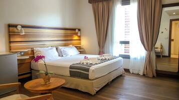 A hotel room with a large bed, bedside tables, a small desk, a chair, a wooden floor, and a window with curtains.
