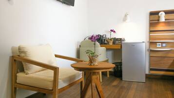 A small kitchenette with a refrigerator, a coffee maker, and a microwave. There is a wooden chair with a cushion and a small wooden table with a vase of flowers.
