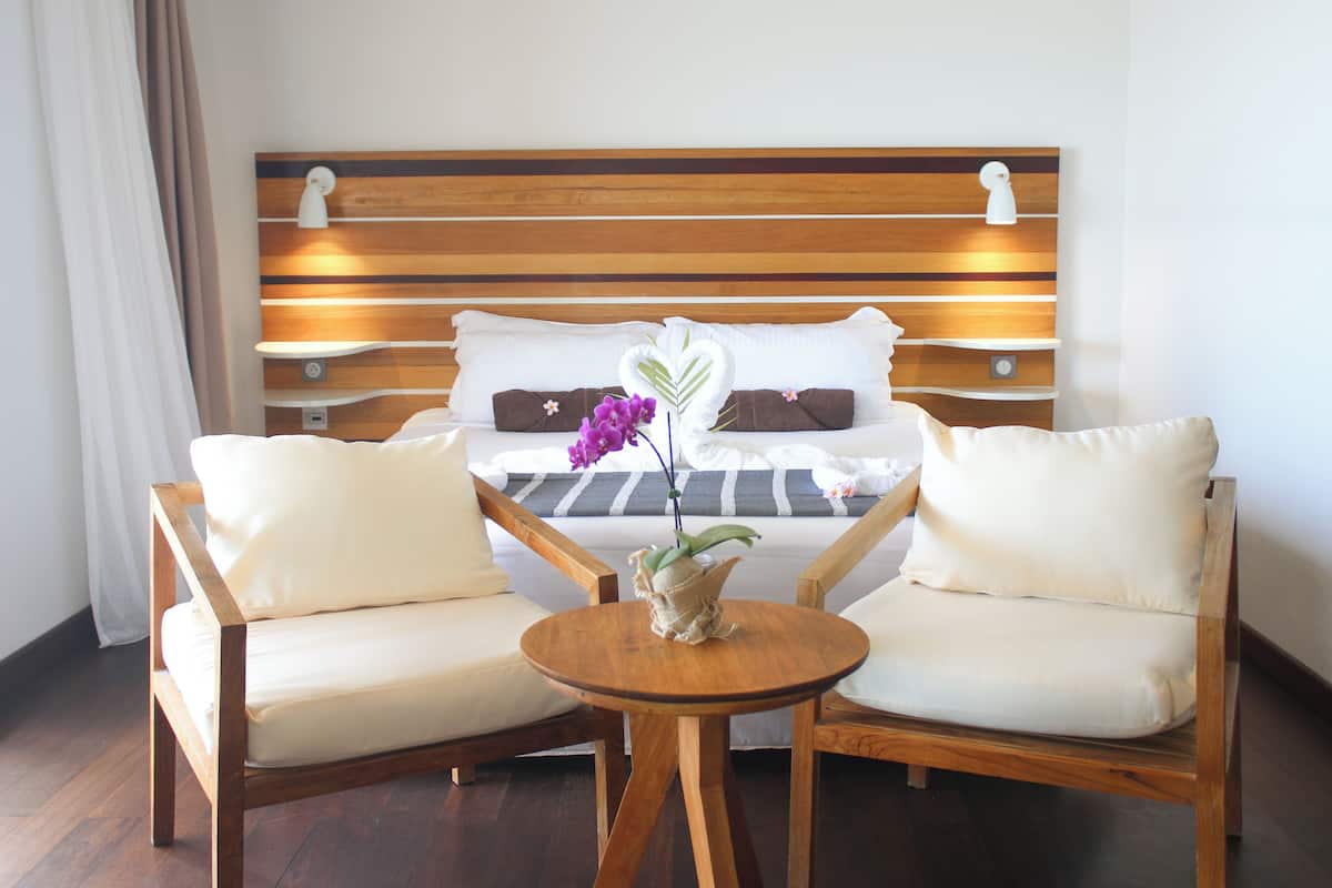 Une chambre d’hôtel avec une tête de lit en bois, deux fauteuils blancs rembourrés, une petite table en bois et un vase rempli de fleurs violettes.
