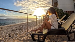 A person sitting on a beach chair overlooking the ocean, with a building and a railing in the background. - Le Battant Des Lames (Saint-Pierre)