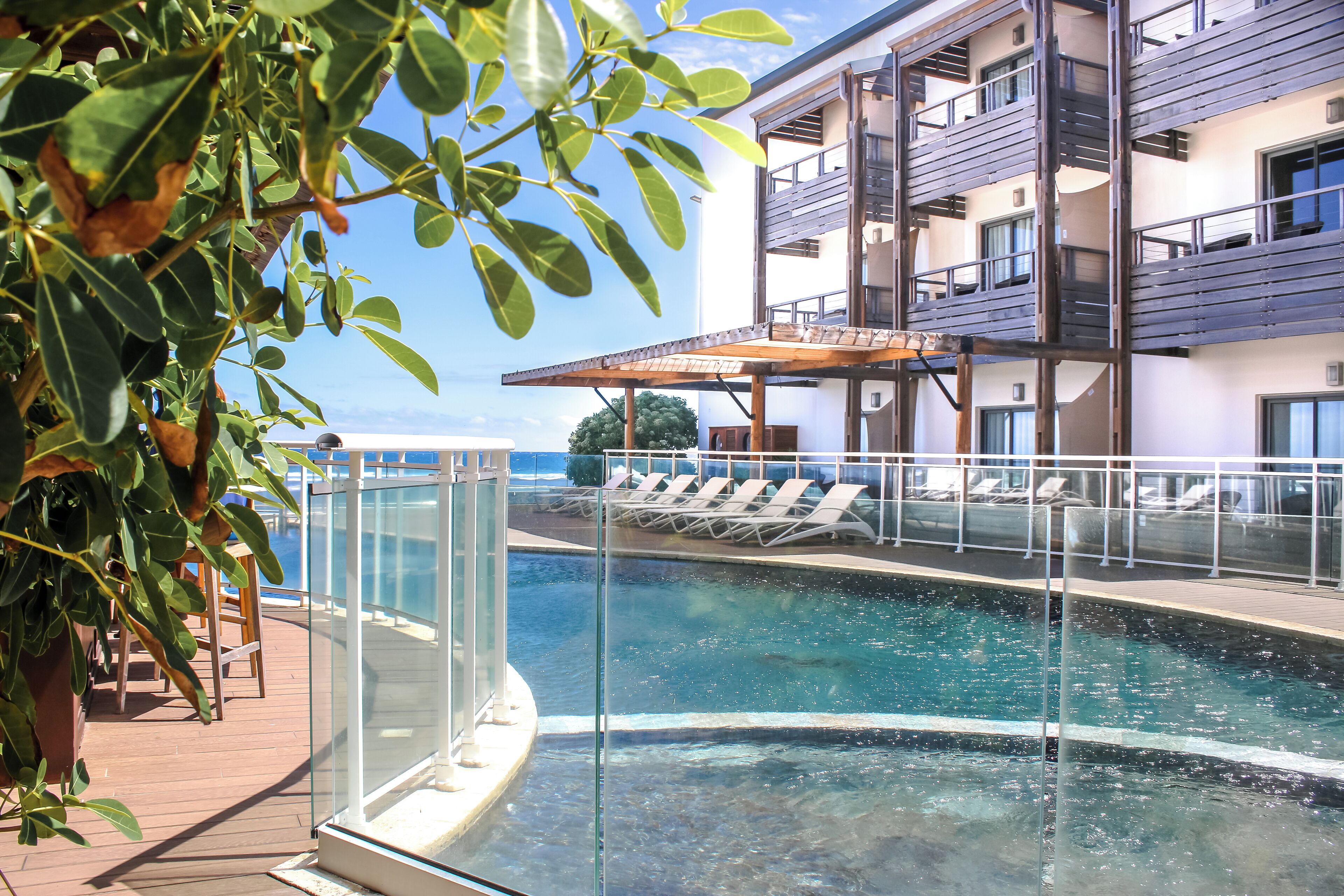 A pool area with a glass barrier, lounge chairs, and a building with balconies.