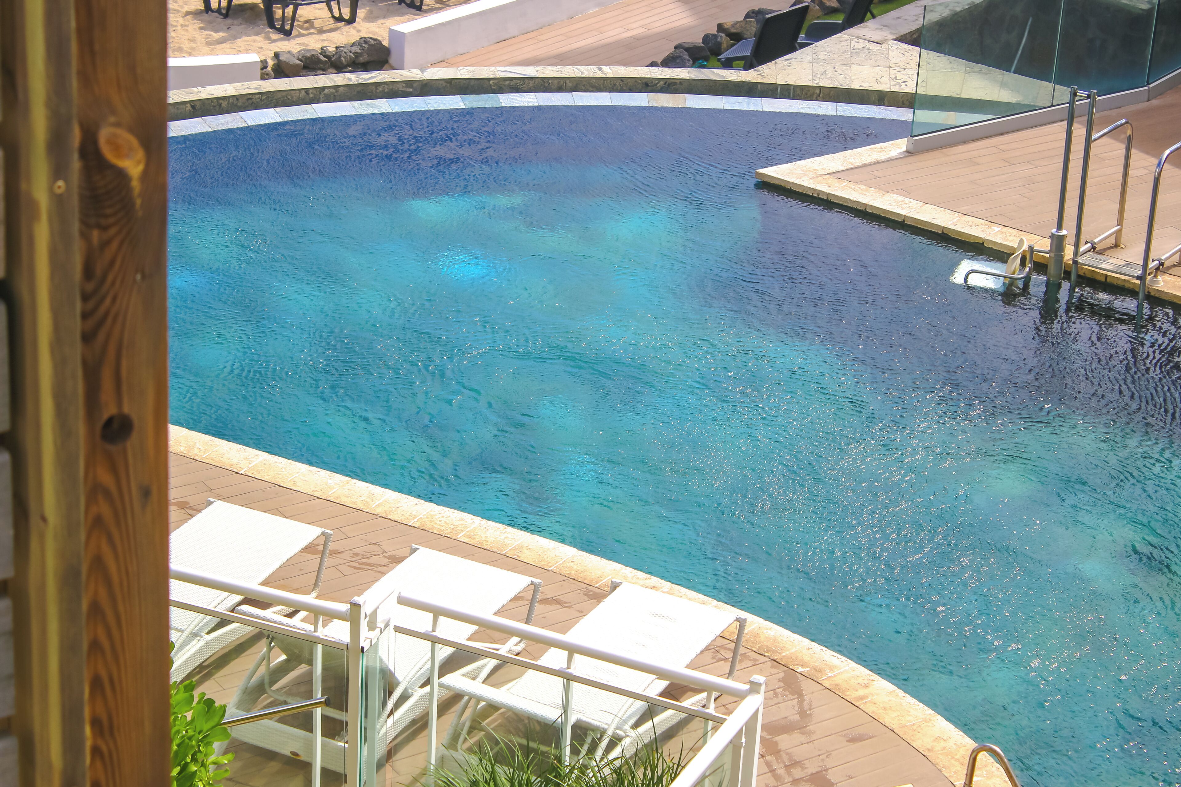 Una piscina con agua cristalina, rodeada por una terraza de madera y sillas reclinables blancas.