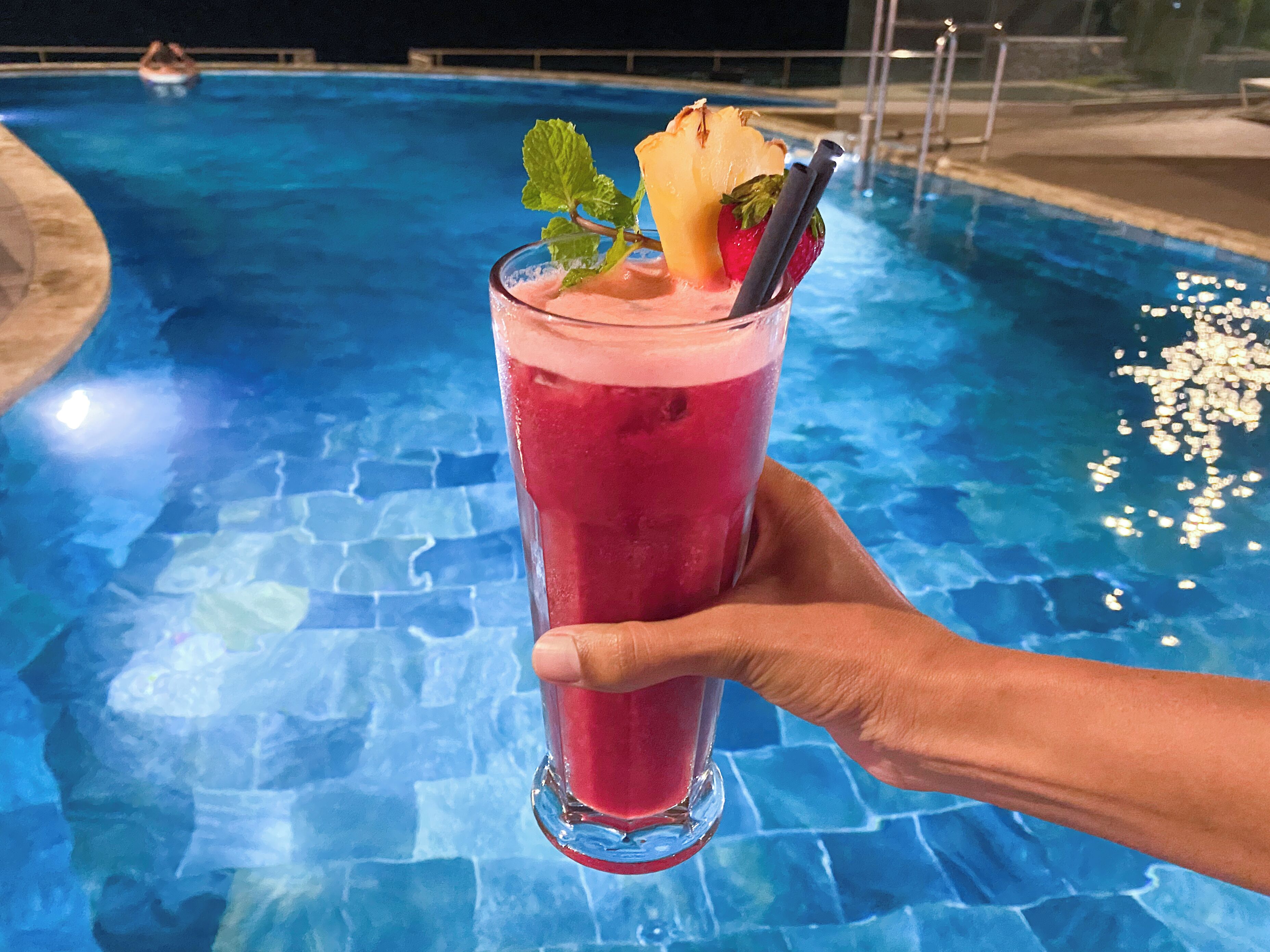Una mano sosteniendo un vaso alto con una bebida rosada, decorado con una rodaja de fruta y una hoja de menta, junto a una piscina.