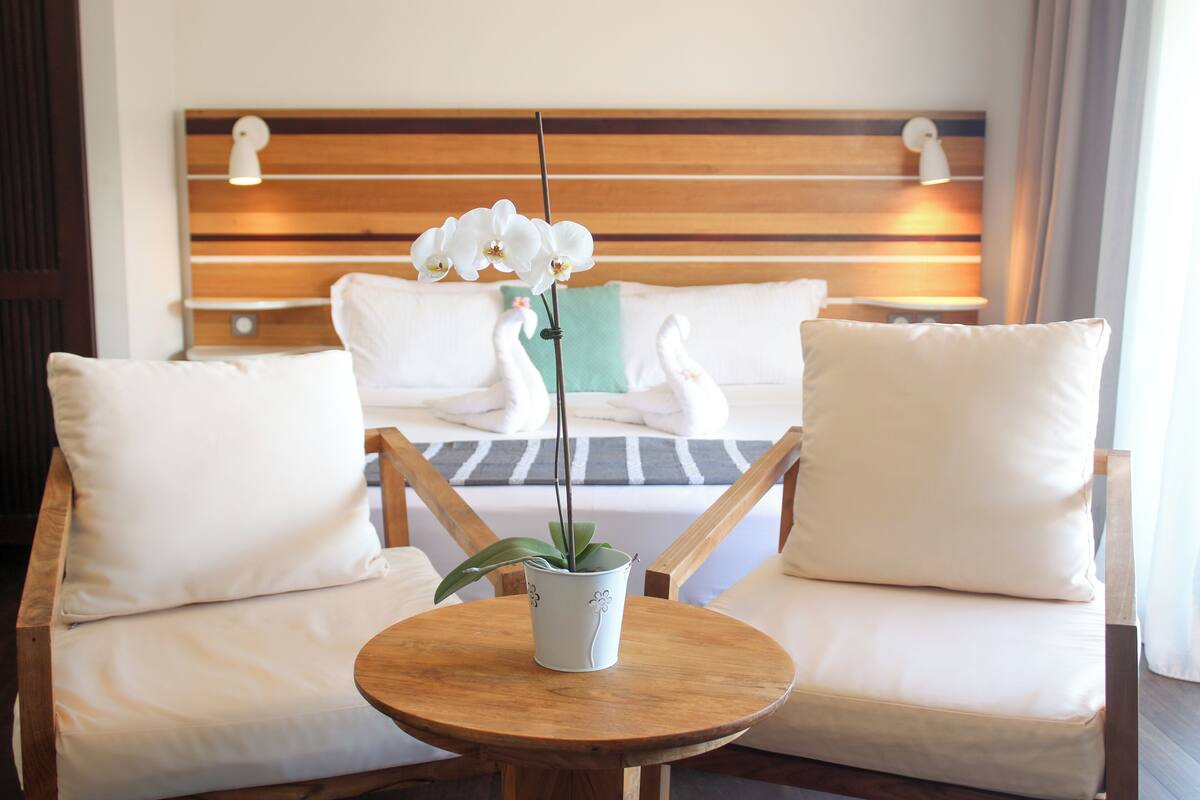 Une chambre d’hôtel avec un lit, deux chaises blanches, une table ronde en bois ornée d’une orchidée en pot, et une tête de lit en bois agrémentée de deux lampes fixées au mur.