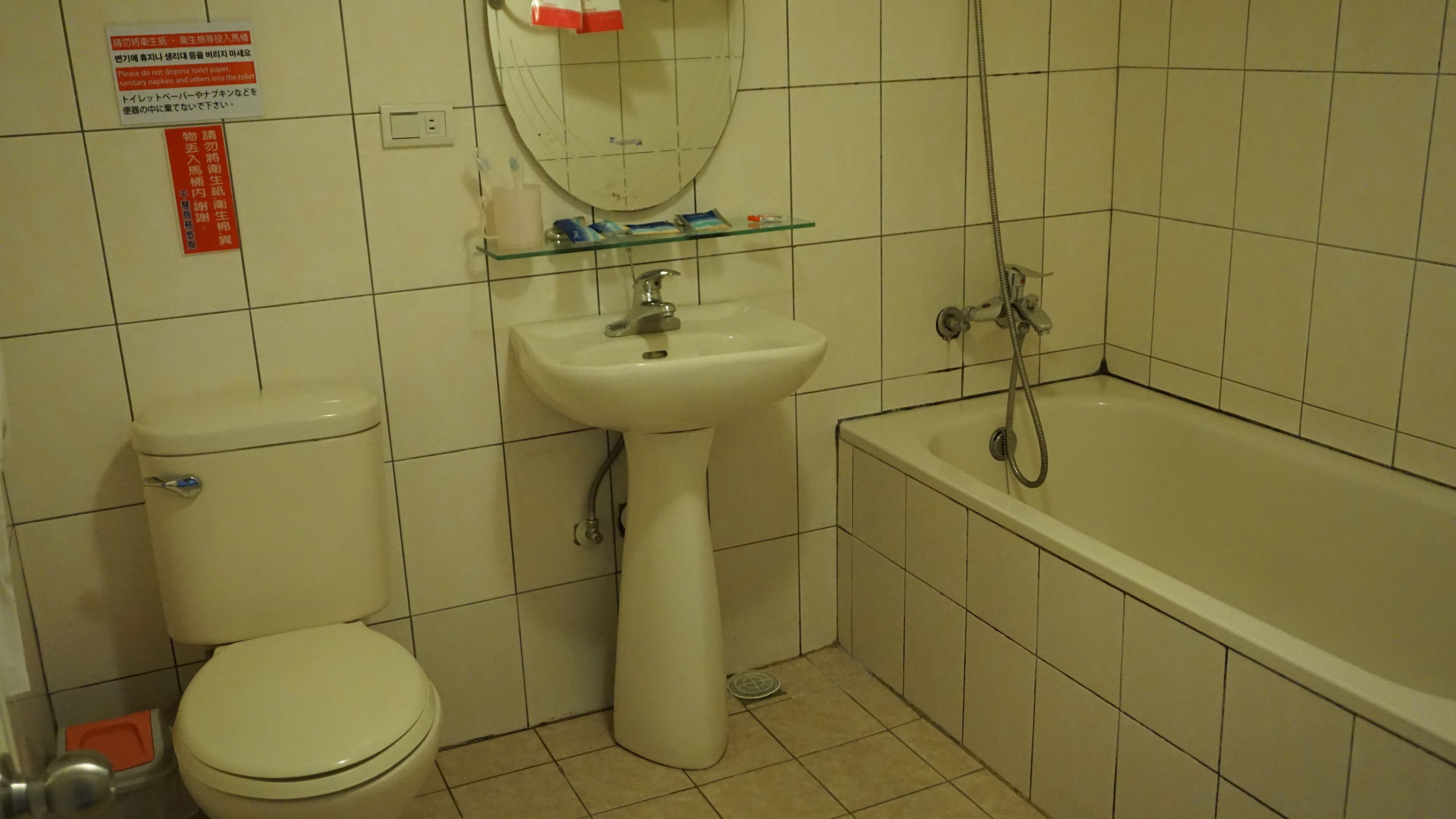 A bathroom with a toilet, sink, and bathtub. There are signs on the wall, a mirror above the sink, and a shelf with toiletries.