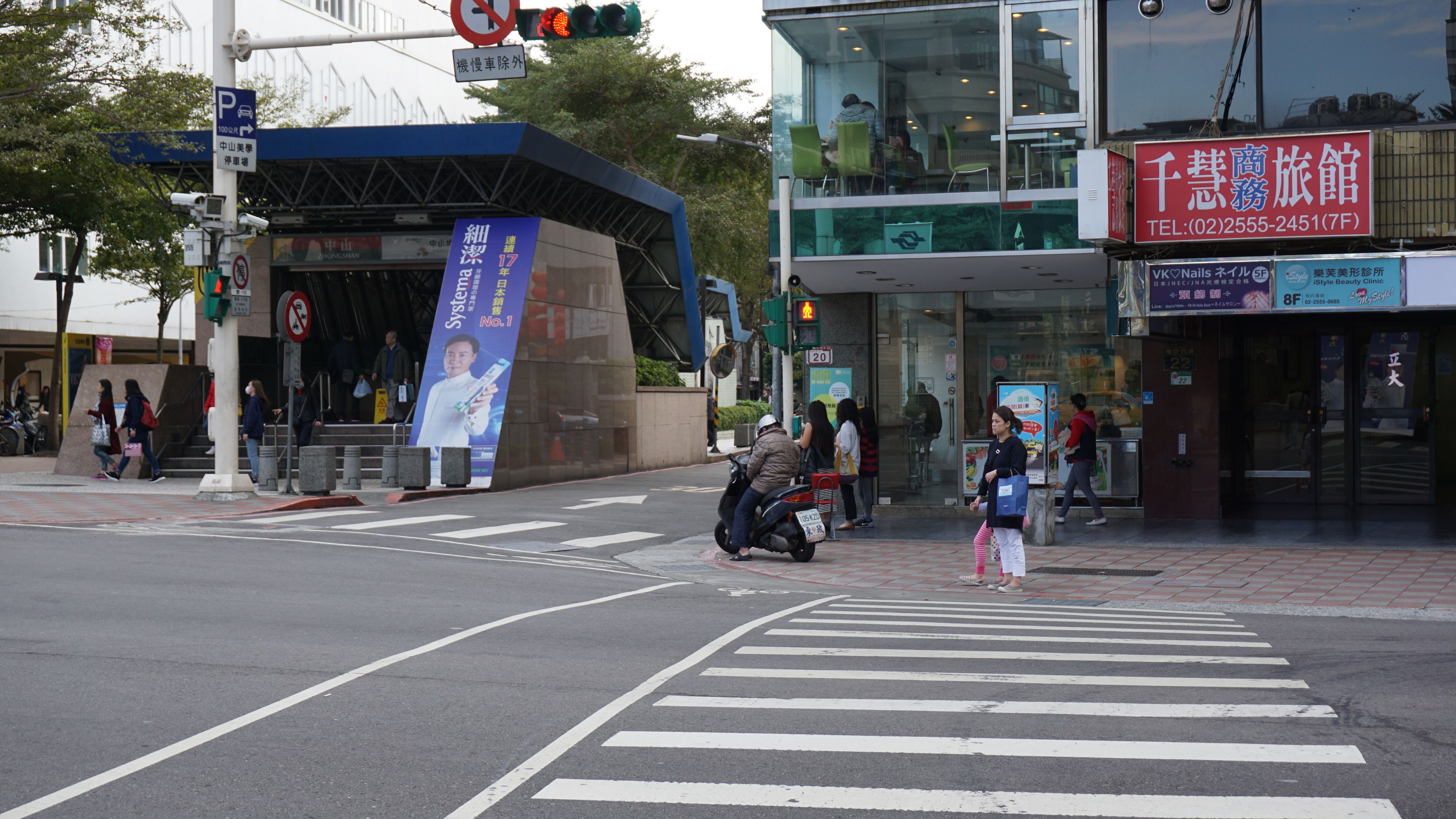 Cruzamento de rua com faixa de pedestres, semáforos e um edifício com placa.