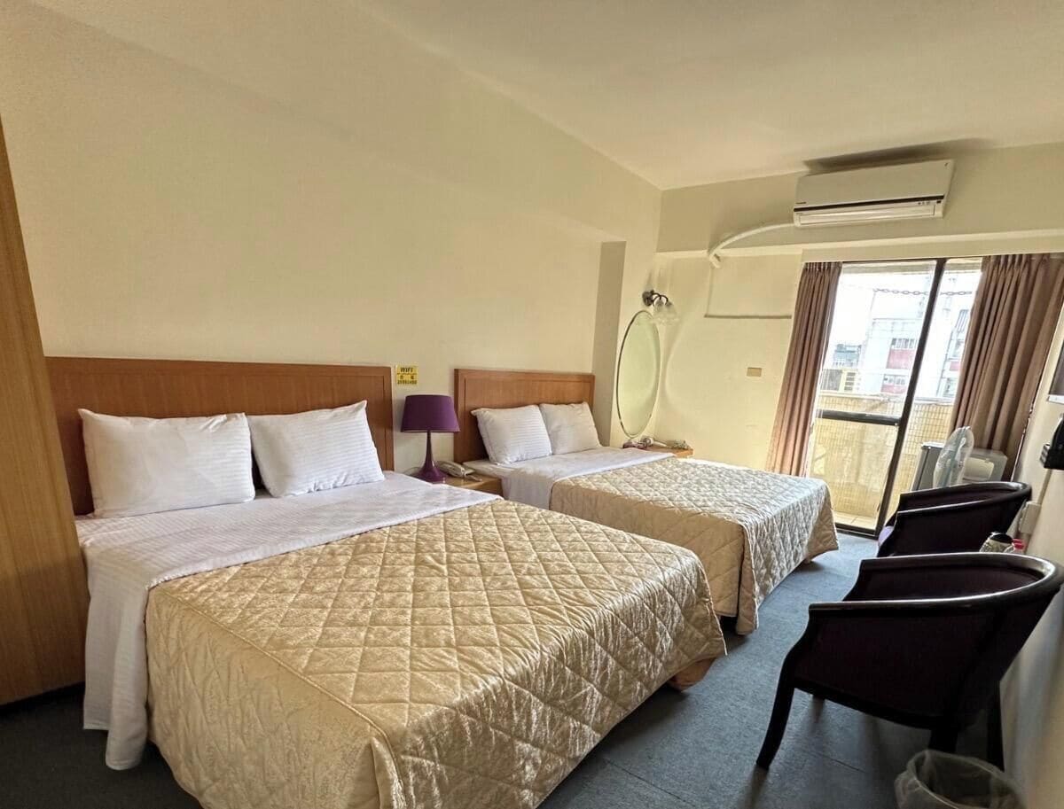 A hotel room with two beds, a desk, a chair, an air conditioning unit, and a view of the outside.