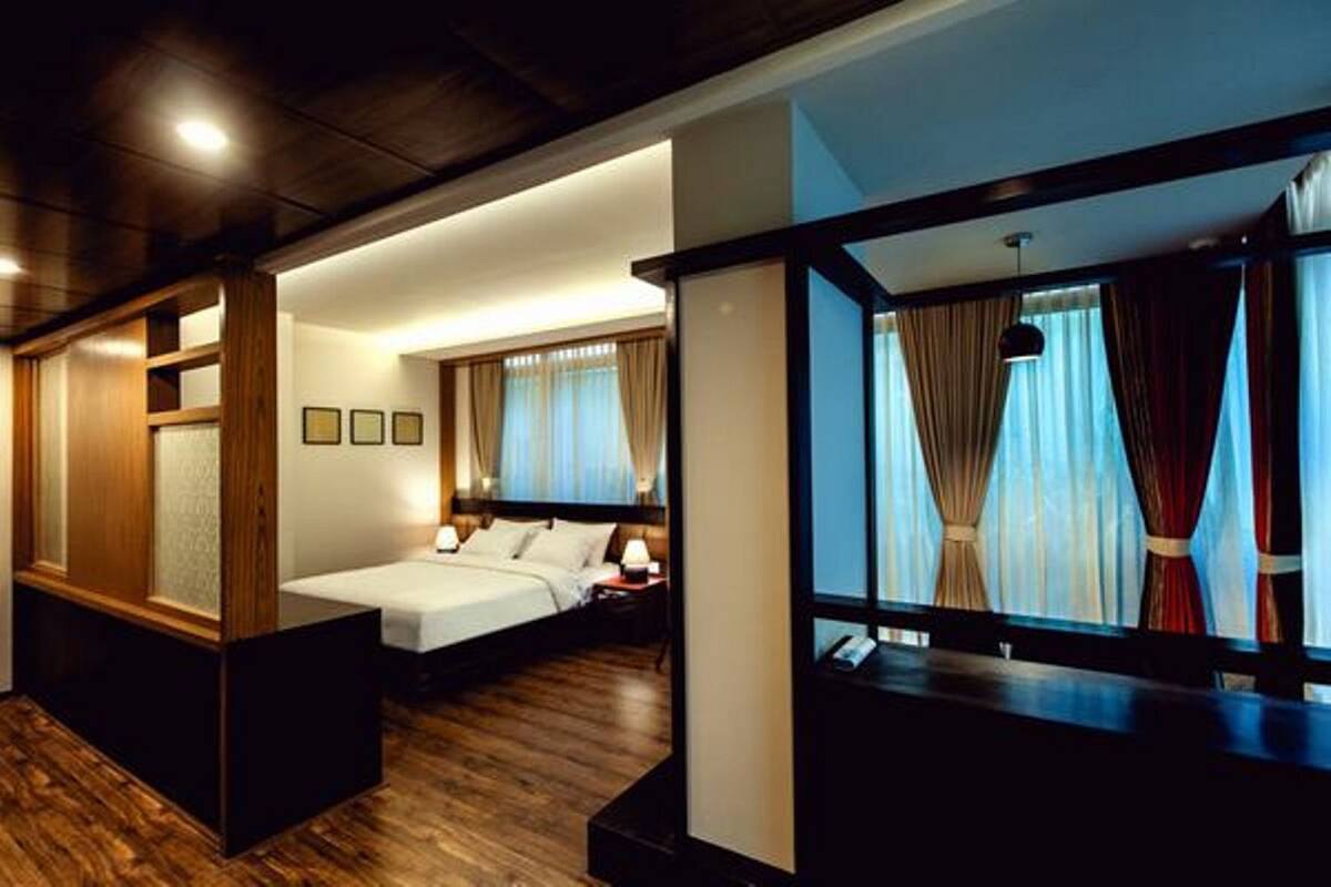 A hotel room with a wooden floor, a bed with white linens, a nightstand with a lamp, and a window with curtains.