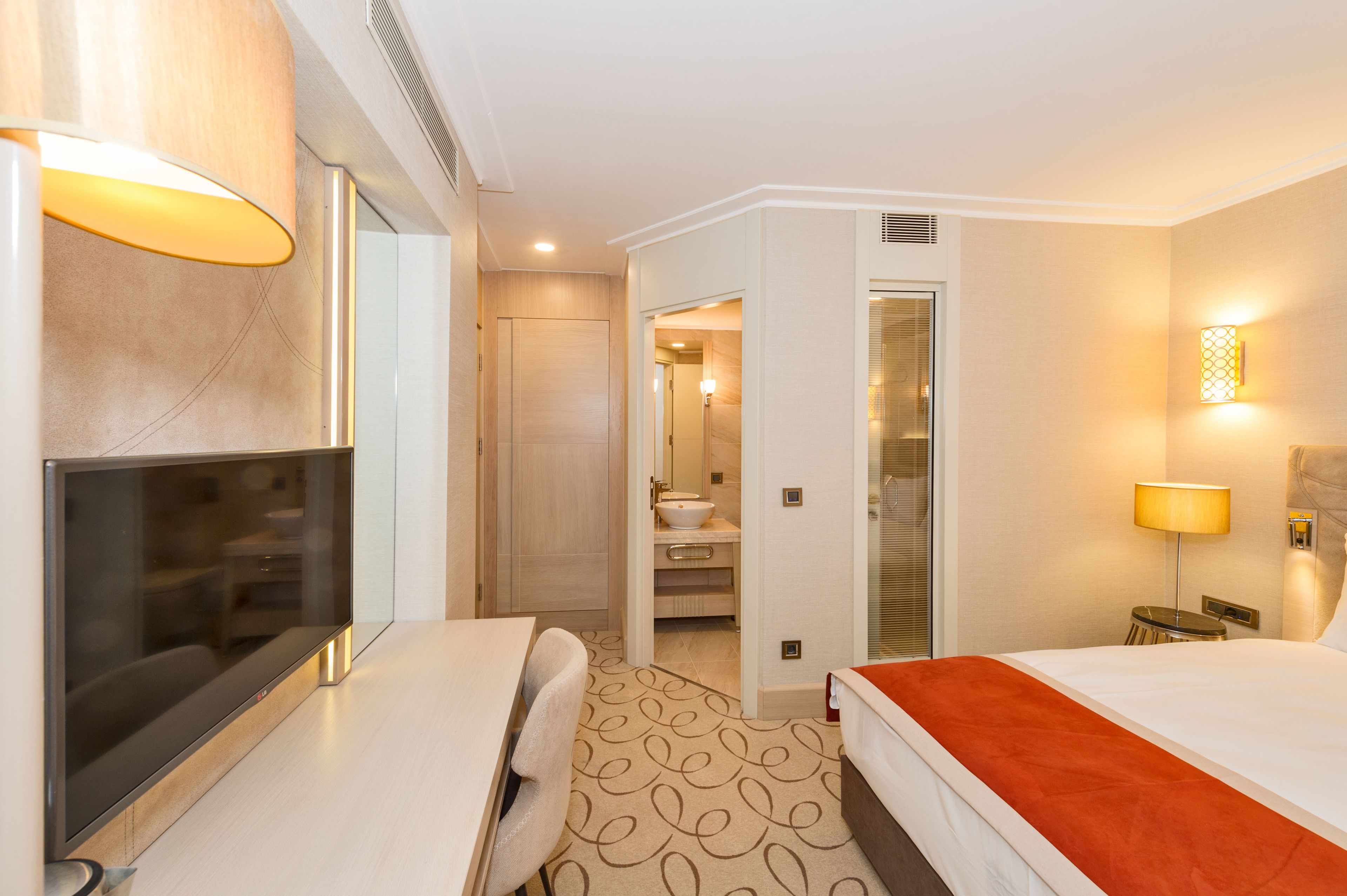 A hotel room with a flat-screen TV, a bed with an orange bedspread, a desk with a chair, and a bathroom visible through an open door.