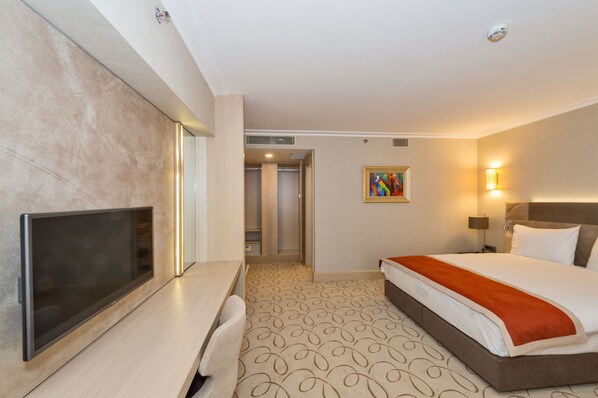 Una habitación de hotel con una televisión de pantalla plana, una cama con una colcha roja, un cuadro en la pared y una alfombra estampada.