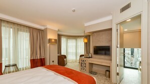Habitación de hotel con una cama grande, un escritorio con televisor de pantalla plana, zona de estar y balcón con vista a la ciudad.