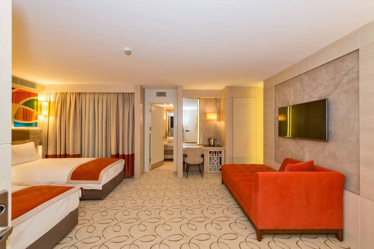 A hotel room with two beds, a sofa, a TV, a desk, and a bathroom visible through an open door.