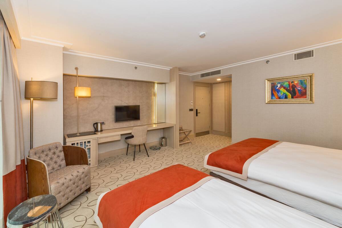 A hotel room with two beds, a desk, a chair, a TV, and a painting on the wall.