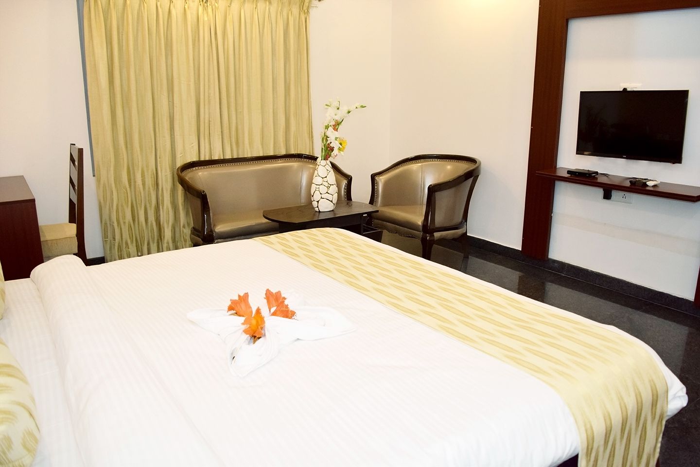Habitación de hotel con una cama grande, dos sillas, una mesita, un florero con flores y un televisor en la pared.