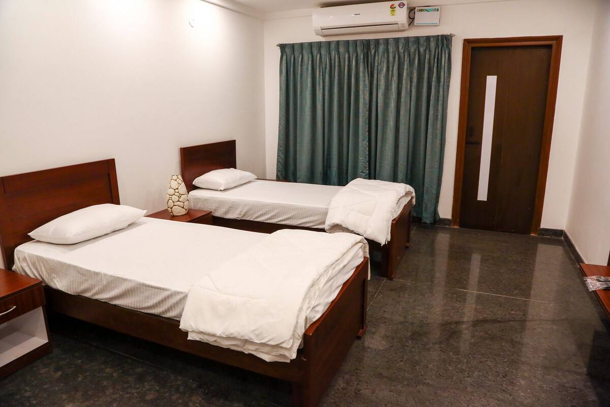 Een hotelkamer met twee eenpersoonsbedden, een nachtkastje met een lamp, een houten deur en een raam met gordijnen.