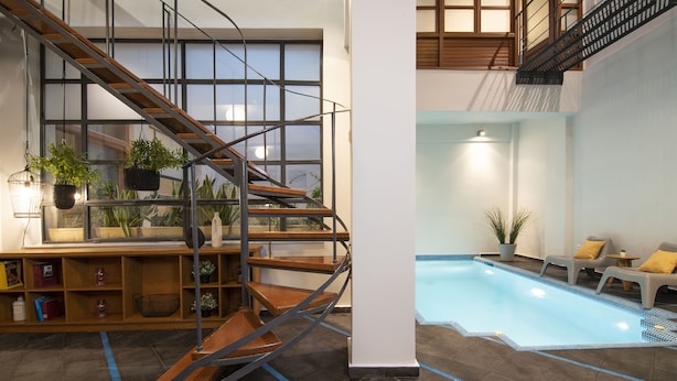 A modern indoor pool area with a wooden spiral staircase, a seating area with cushions, and a bookshelf with plants. - Villa Meliti (Athens)