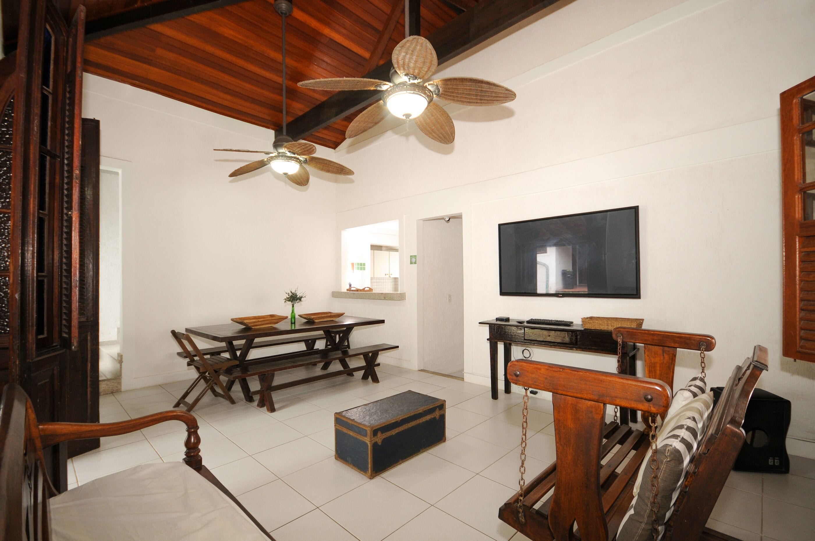 Amplia sala de estar con techo de madera, una mesa de comedor con bancos, televisor montado en la pared y un gran ventanal.