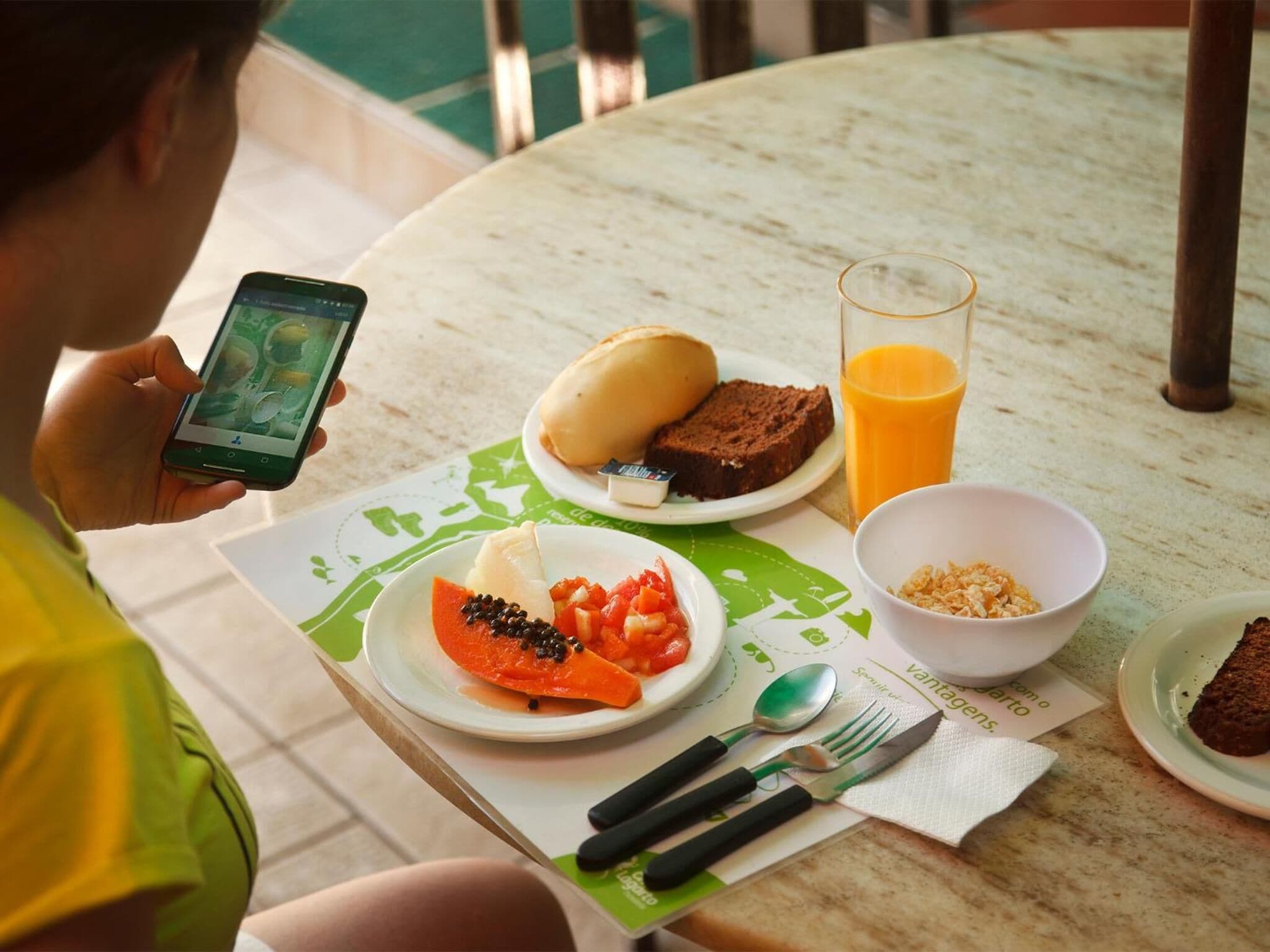 A person at a table with a meal consisting of a sandwich, a glass of orange juice, a bowl of cereal, and a plate with fruit and a dessert.