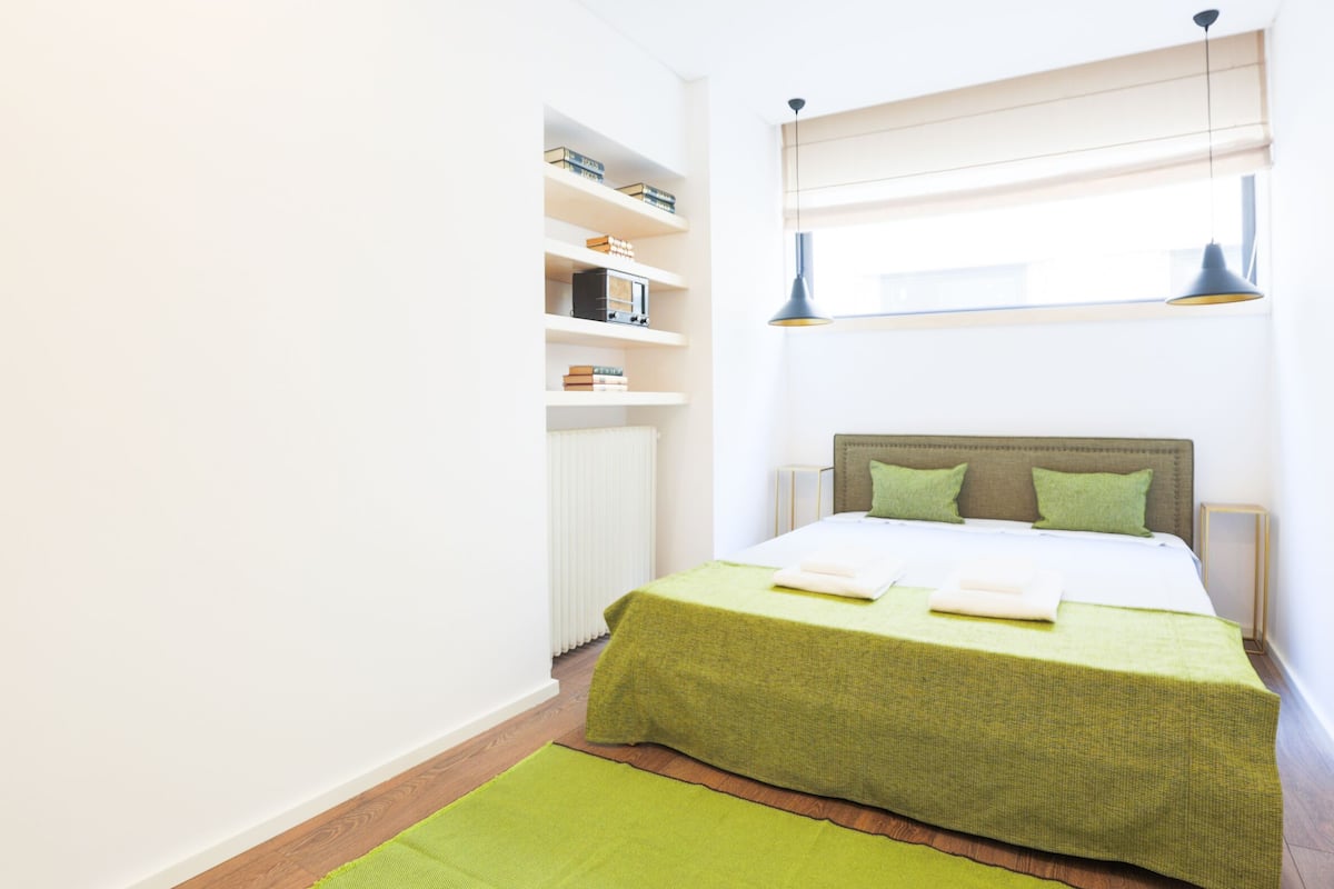A neatly arranged bedroom with a bed, green bedding, a bookshelf, and a window with blinds.