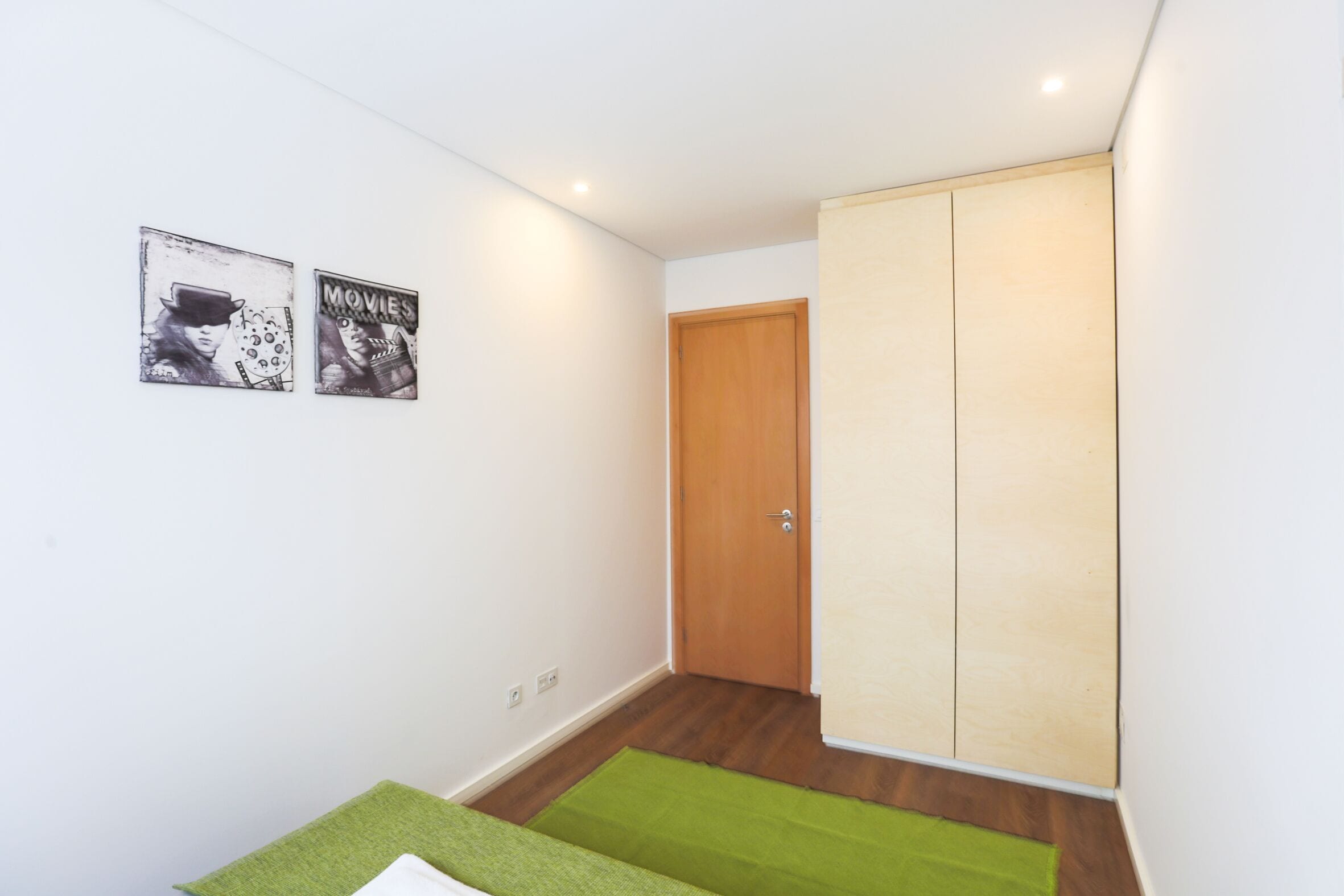 A room with a bed, a green rug, a wooden door, and two posters on the wall.