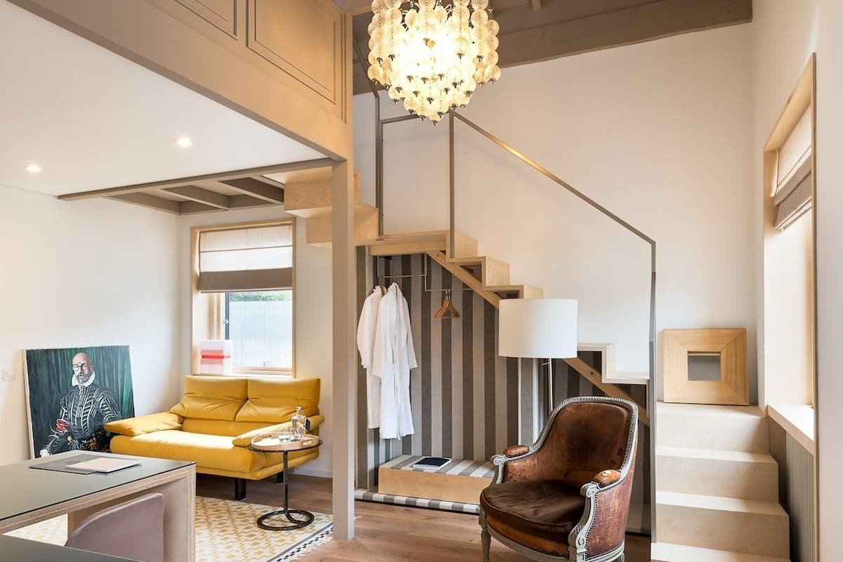 A spacious living area with a staircase, a chandelier, a yellow sofa, and a leather armchair.
