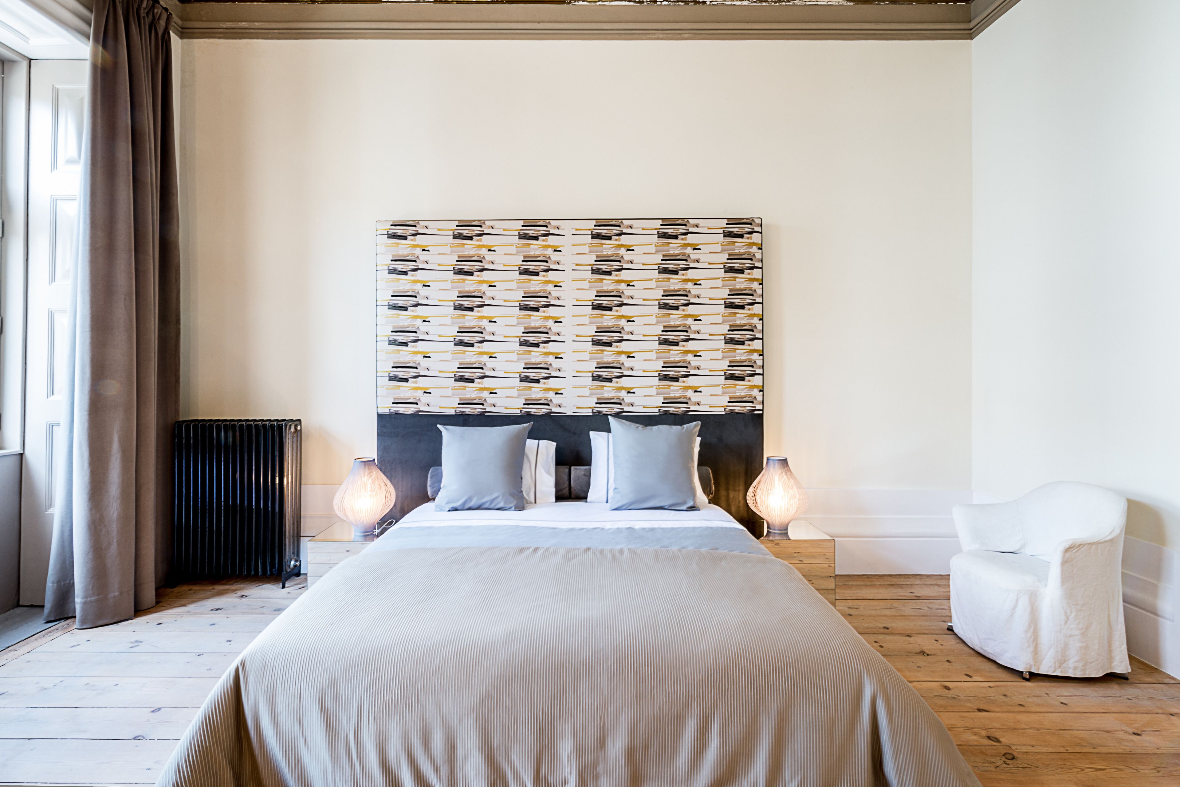 Un dormitorio con una cama grande, cabecera con diseño estampado, dos lámparas de noche, un radiador y una silla blanca.