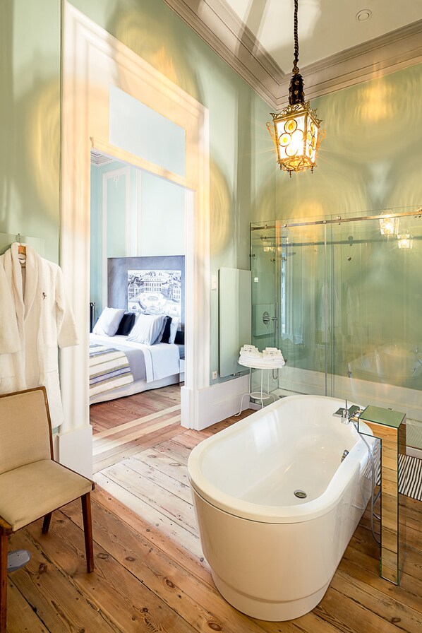 A modern bathroom with a freestanding bathtub, a glass shower enclosure, and a visible bedroom with a bed and a large window.