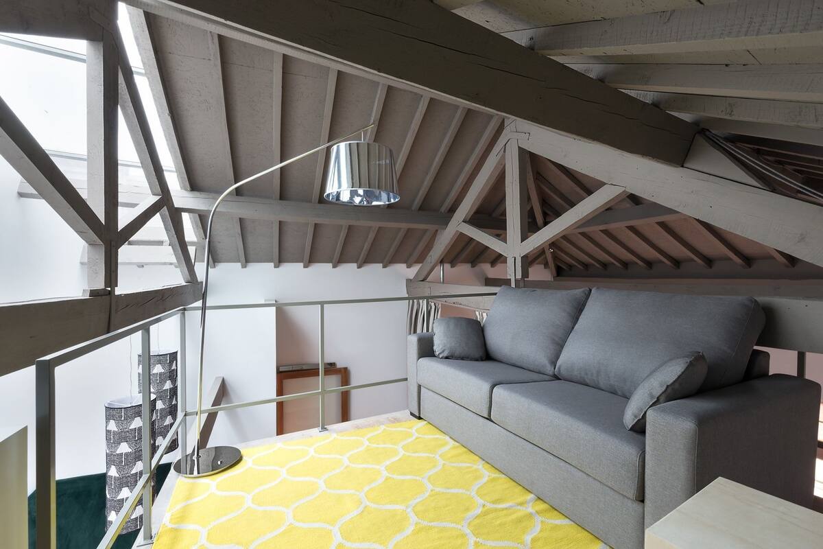 A loft-style room with exposed wooden beams, a gray sofa, and a yellow patterned rug.