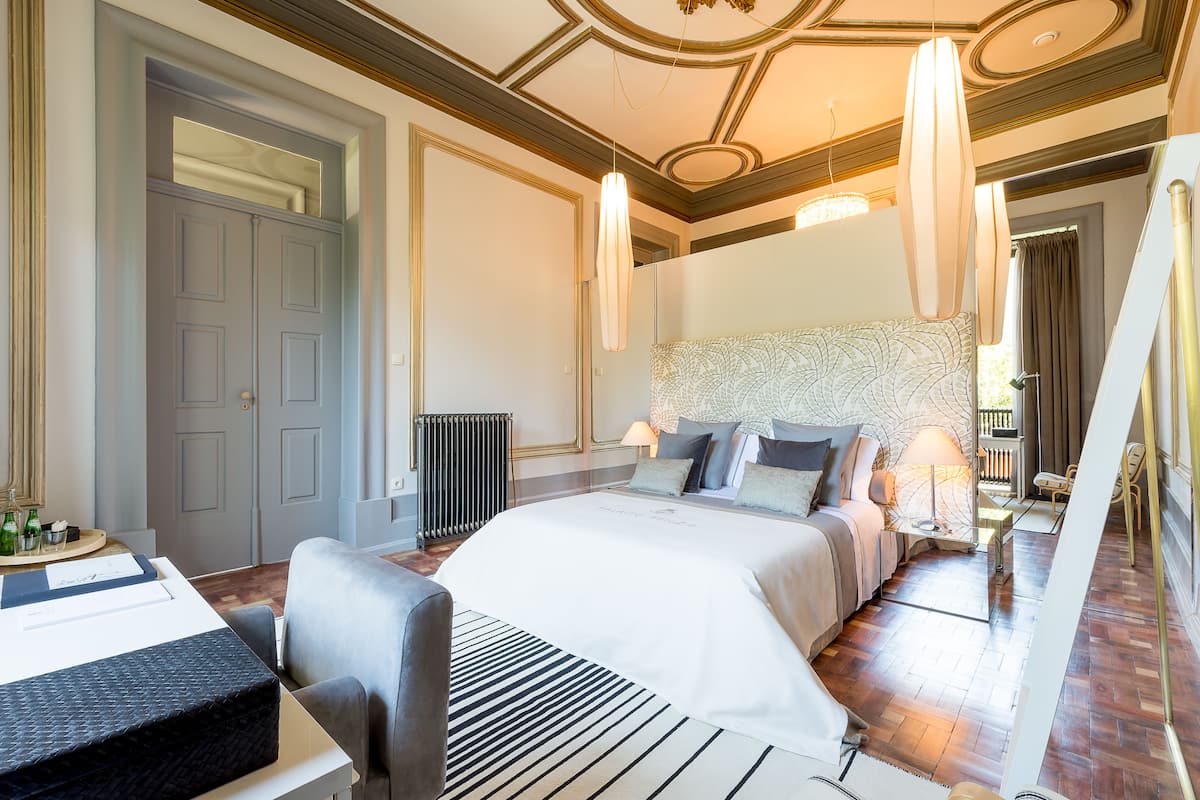 A hotel room with a large bed, a desk, and a chair. There is a radiator on the wall and a window with curtains.