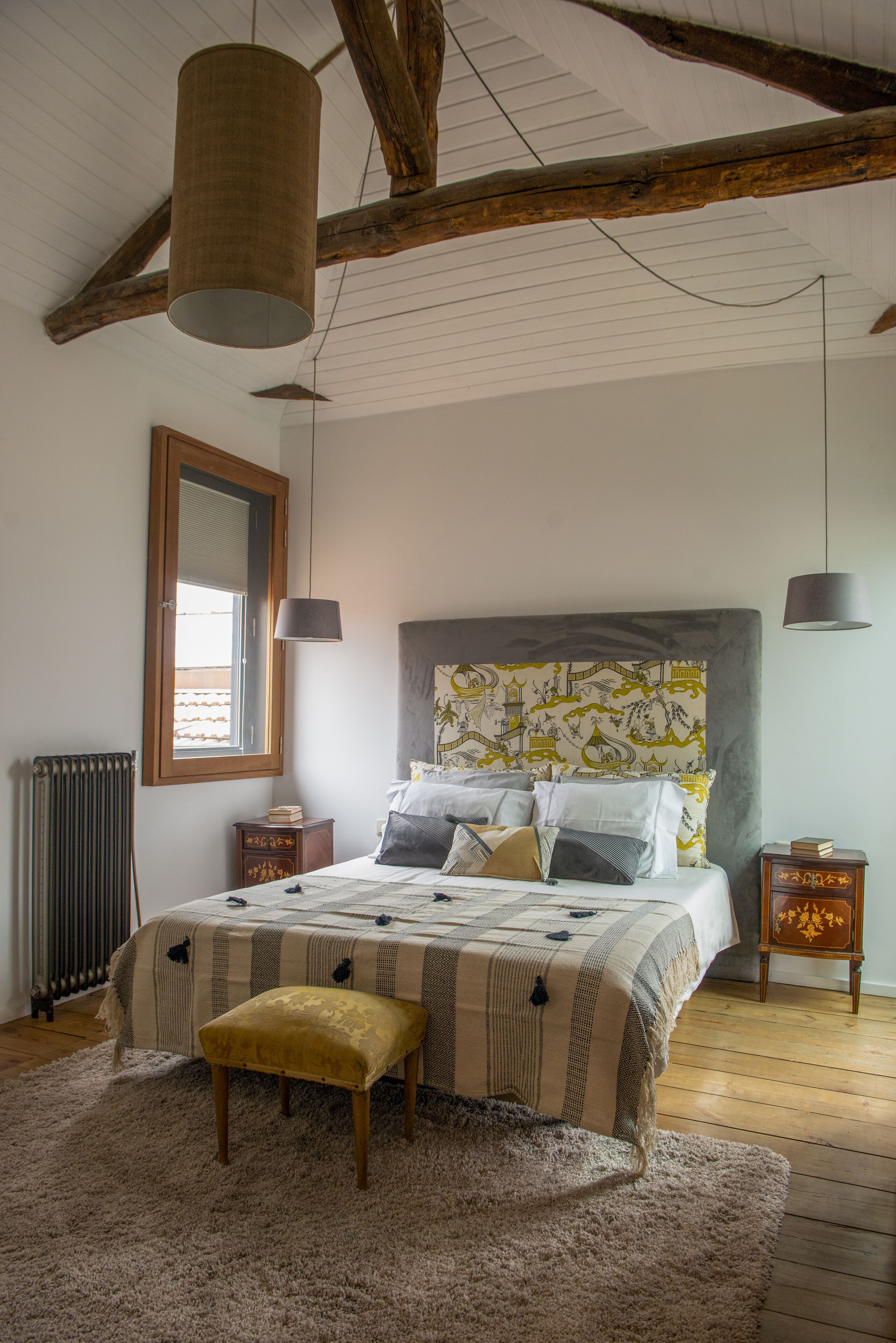 Un dormitorio con una cama grande, un cabecero de madera, una ventana, un radiador y una mesita de noche pequeña de madera.