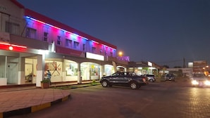 A street view at dusk with illuminated storefronts, parked cars, and a pedestrian walkway. - Hotel Al-Khalil Machava (Matola)