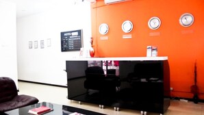 A reception area with a black desk, a red chair, and a glass table. There are clocks on the wall and a sign with room rates. - Hotel Al-Khalil Machava (Matola)