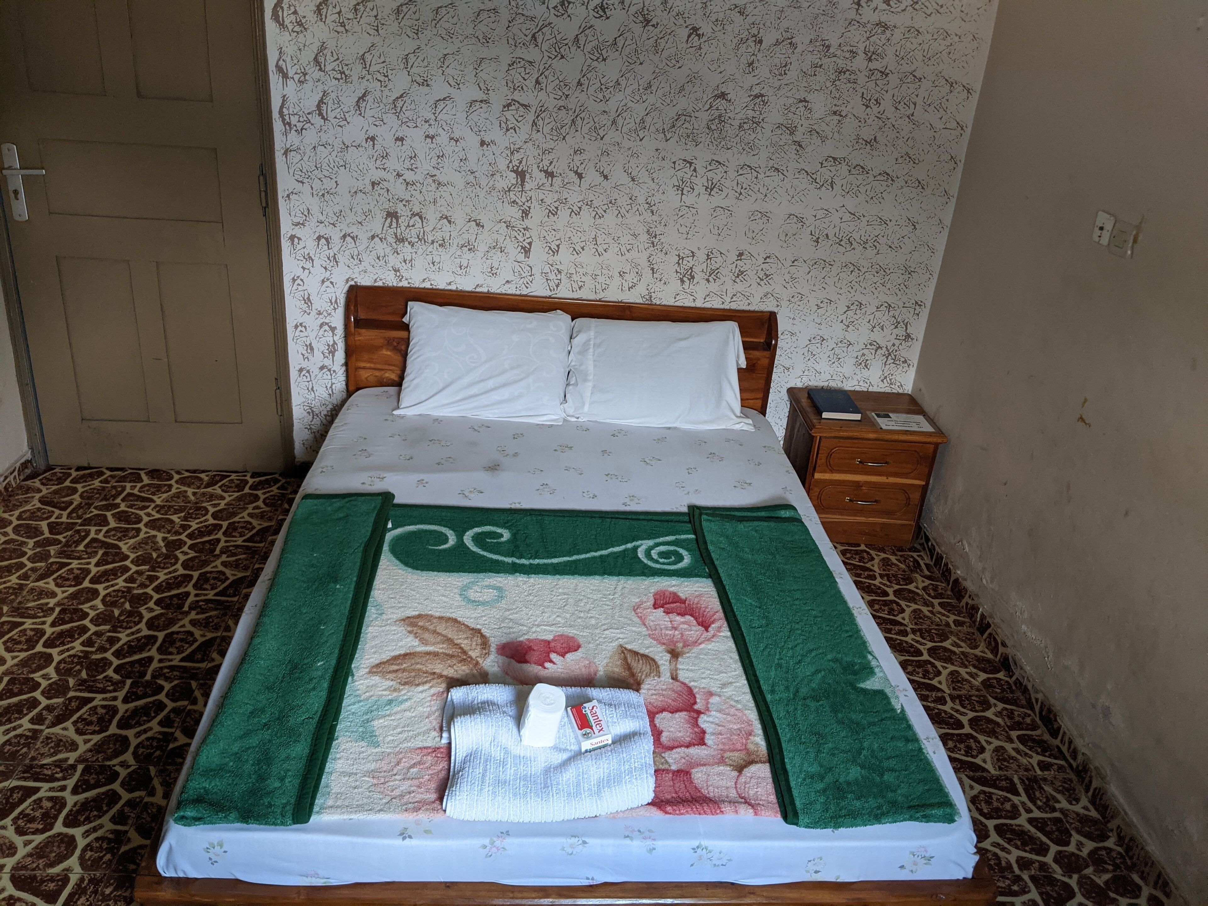 Un lit bien fait, recouvert d’une couvre-lit à motifs floraux et agrémenté de serviettes vertes, une table de chevet en bois avec un livre, et un sol à motifs.