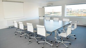 Un salón de conferencias con una gran mesa, sillas blancas, una pizarra y ventanas amplias con vista a edificios.