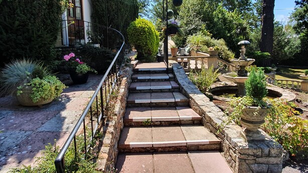 Una escalera de piedra conduce a un espacio cubierto con un dosel blanco, rodeado de exuberante vegetación y una pequeña fuente.