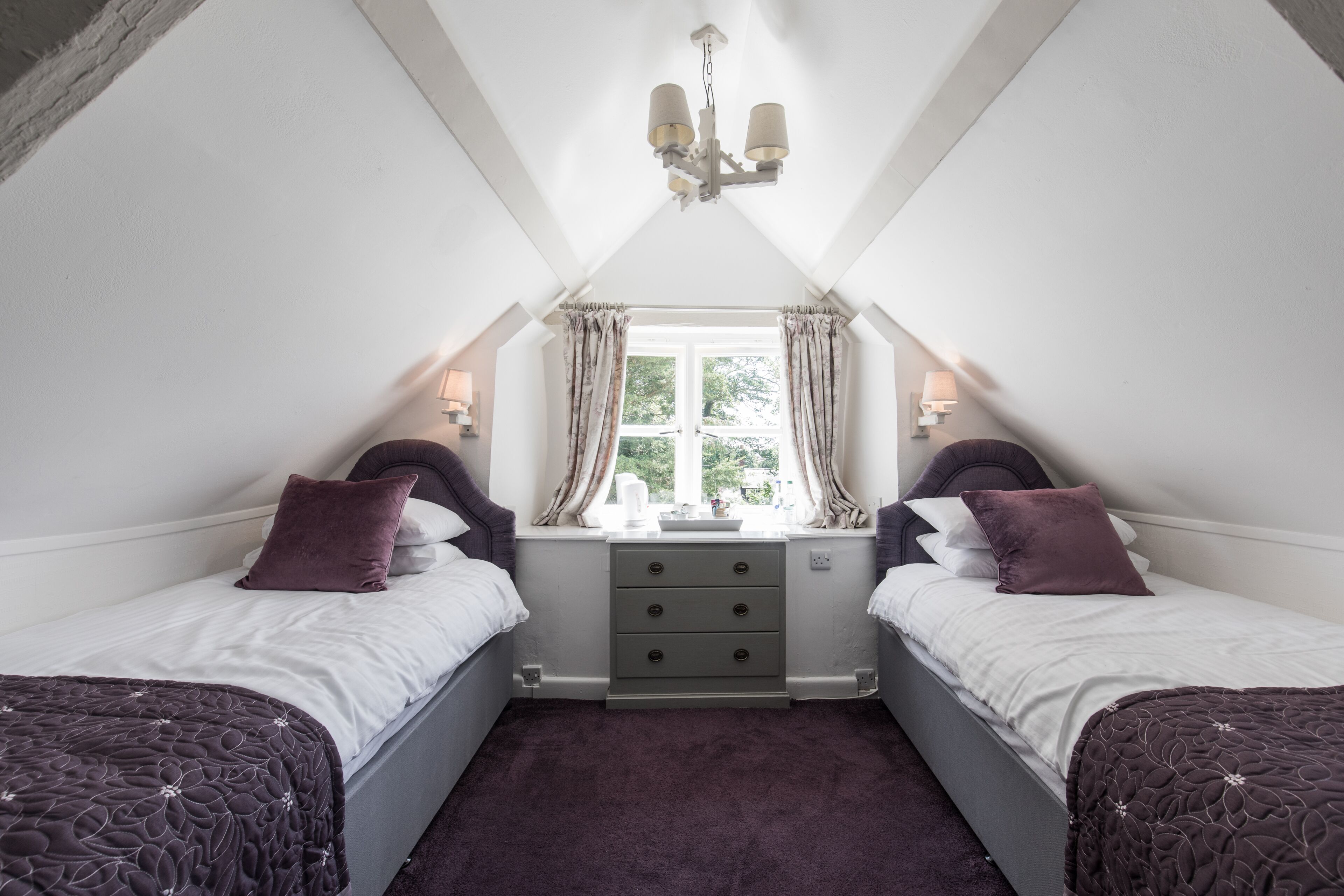A bedroom with two single beds, a window with curtains, a bedside table with lamps, and a chandelier.