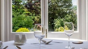 Mesa para dos con mantel blanco, cubiertos y copas de vino, con vista a un jardín exuberante.