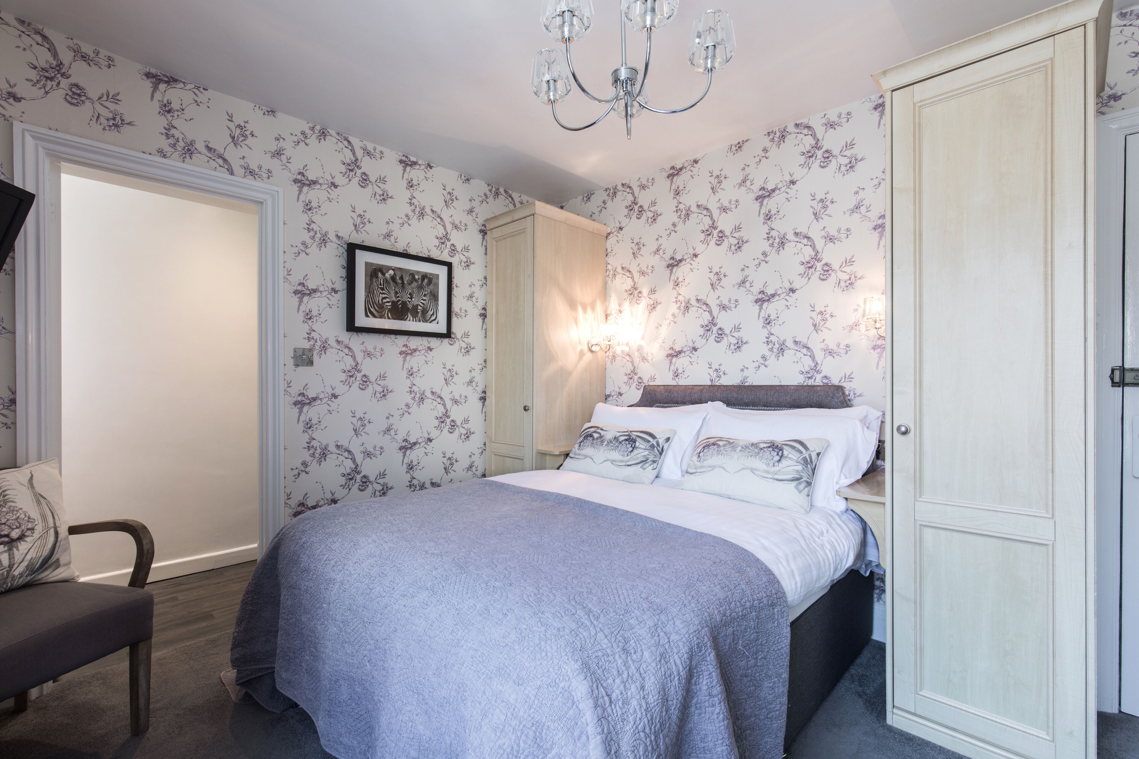 A bedroom with a bed, a chair, a wardrobe, and a wall-mounted light fixture.